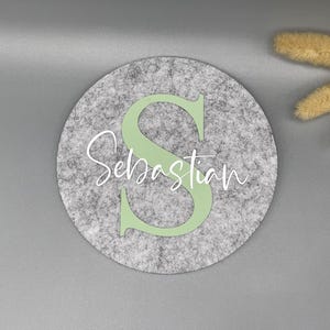 Puede incluir: Decoraci&oacute;n redonda de fieltro gris con el nombre "Sebastian" en escritura blanca y una gran "S" verde claro. El fieltro tiene una textura moteada. Flores secas en la esquina superior derecha.