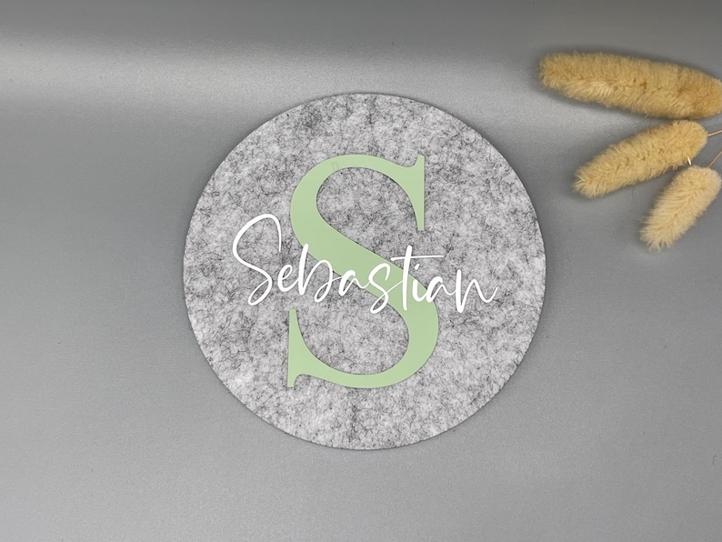 Puede incluir: Decoraci&oacute;n redonda de fieltro gris con el nombre "Sebastian" en escritura blanca y una gran "S" verde claro. El fieltro tiene una textura moteada. Flores secas en la esquina superior derecha.