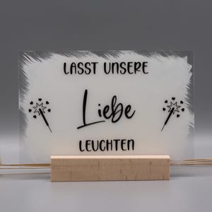 May include: A rectangular clear acrylic sign with a white painted background, featuring the black text "LASST UNSERE Liebe LEUCHTEN" and two black sparkler icons with small hearts. The sign is held upright by a light-colored wooden block base.