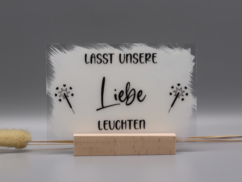 May include: A rectangular clear acrylic sign with a white painted background, featuring the black text "LASST UNSERE Liebe LEUCHTEN" and two black sparkler icons with small hearts. The sign is held upright by a light-colored wooden block base.
