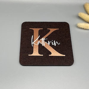 Puede incluir: Un posavasos cuadrado marr&oacute;n oscuro con la letra "K" en color cobre y el nombre "Kathrin" en escritura blanca. El posavasos est&aacute; hecho de un material similar al fieltro. Flores secas decorativas en el fondo.
