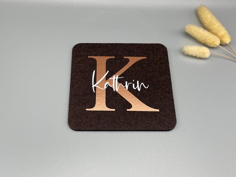 Puede incluir: Un posavasos cuadrado marr&oacute;n oscuro con la letra "K" en color cobre y el nombre "Kathrin" en escritura blanca. El posavasos est&aacute; hecho de un material similar al fieltro. Flores secas decorativas en el fondo.