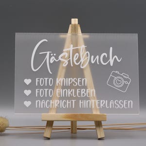 May include: A rectangular frosted acrylic sign displayed on a small wooden easel. The sign features white text reading "G&auml;stebuch," "Foto knipsen," "Foto einkleben," and "Nachricht hinterlassen," accompanied by three heart icons and a camera icon.