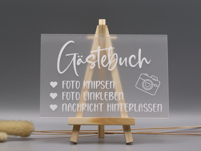 May include: A rectangular frosted acrylic sign displayed on a small wooden easel. The sign features white text reading "G&auml;stebuch," "Foto knipsen," "Foto einkleben," and "Nachricht hinterlassen," accompanied by three heart icons and a camera icon.