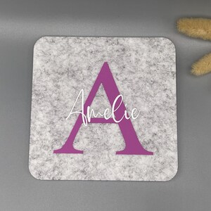 Puede incluir: Un posavasos cuadrado de fieltro gris con una gran letra "A" morada y el nombre "Amelie" en escritura blanca. El posavasos tiene esquinas redondeadas y un aspecto texturizado. Flores secas decorativas en el fondo.