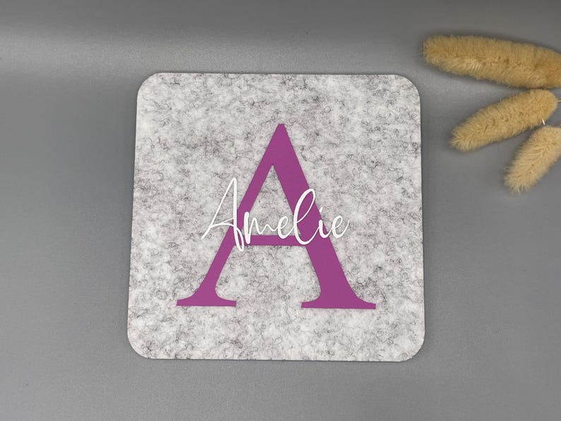 Puede incluir: Un posavasos cuadrado de fieltro gris con una gran letra "A" morada y el nombre "Amelie" en escritura blanca. El posavasos tiene esquinas redondeadas y un aspecto texturizado. Flores secas decorativas en el fondo.