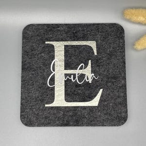 Puede incluir: Un posavasos cuadrado de fieltro gris oscuro con una gran letra "E" blanca y el nombre "Emilia" en una escritura delicada. El posavasos es un dise&ntilde;o simple y elegante.