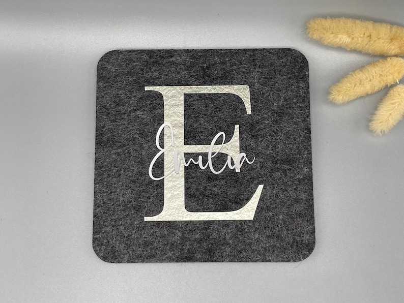 Puede incluir: Un posavasos cuadrado de fieltro gris oscuro con una gran letra "E" blanca y el nombre "Emilia" en una escritura delicada. El posavasos es un dise&ntilde;o simple y elegante.