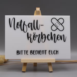 May include: A rectangular white sign resting on a small wooden easel. The sign features black text that reads Notfall-K&ouml;rbchen and BITTE BEDIENT EUCH, accompanied by a black graphic of two crossed adhesive bandages with small heart details.