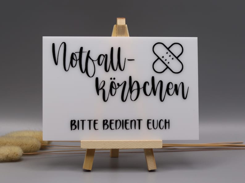 May include: A rectangular white sign resting on a small wooden easel. The sign features black text that reads Notfall-K&ouml;rbchen and BITTE BEDIENT EUCH, accompanied by a black graphic of two crossed adhesive bandages with small heart details.