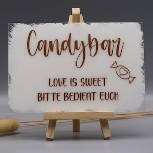 May include: A rectangular frosted acrylic sign with white painted edges, displayed on a small wooden easel. The sign features brown text reading Candybar, LOVE IS SWEET, and BITTE BEDIENT EUCH, along with a small graphic of a wrapped candy with a heart.