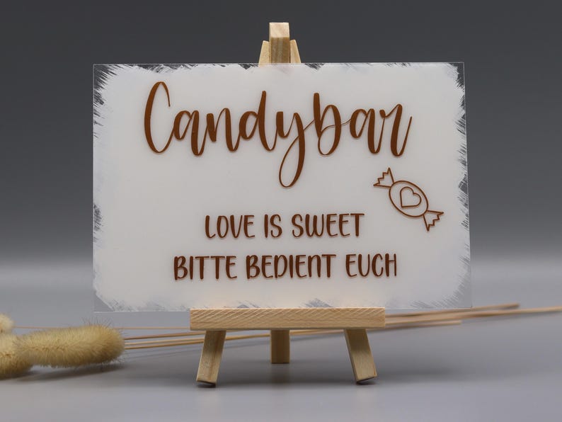 May include: A rectangular frosted acrylic sign with white painted edges, displayed on a small wooden easel. The sign features brown text reading Candybar, LOVE IS SWEET, and BITTE BEDIENT EUCH, along with a small graphic of a wrapped candy with a heart.