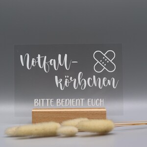 May include: A rectangular clear acrylic sign with white text reading Notfall-K&ouml;rbchen and BITTE BEDIENT EUCH, featuring a white graphic of two crossed adhesive bandages, set into a light-colored wooden base with dried beige grass stems in front.