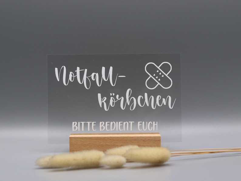 May include: A rectangular clear acrylic sign with white text reading Notfall-K&ouml;rbchen and BITTE BEDIENT EUCH, featuring a white graphic of two crossed adhesive bandages, set into a light-colored wooden base with dried beige grass stems in front.