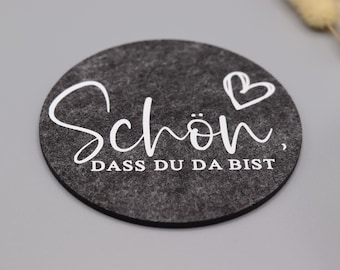 May include: A circular dark gray felt coaster featuring white printed text that reads "Schön, DASS DU DA BIST" in a combination of cursive and block lettering, accompanied by a small hand-drawn heart outline above the word "Schön".
