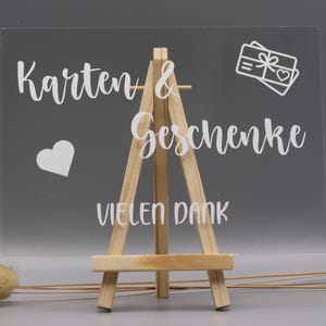 May include: A clear rectangular acrylic sign displayed on a small wooden easel. The sign features white text that reads "Karten & Geschenke" and "VIELEN DANK," accompanied by a white graphic of a gift box with a heart and a separate heart icon.