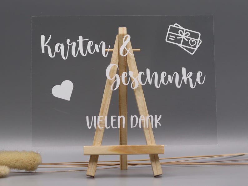 May include: A clear rectangular acrylic sign displayed on a small wooden easel. The sign features white text that reads "Karten & Geschenke" and "VIELEN DANK," accompanied by a white graphic of a gift box with a heart and a separate heart icon.