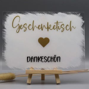 May include: A rectangular clear acrylic sign featuring a white painted brushstroke background. The text "Geschenktisch" is in gold script, followed by a gold heart icon and the word "DANKESCH&Ouml;N" in black sans-serif font, displayed on a small wooden easel.