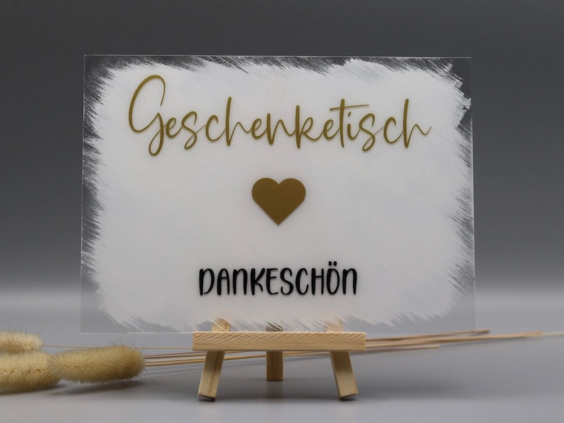 May include: A rectangular clear acrylic sign featuring a white painted brushstroke background. The text "Geschenktisch" is in gold script, followed by a gold heart icon and the word "DANKESCH&Ouml;N" in black sans-serif font, displayed on a small wooden easel.