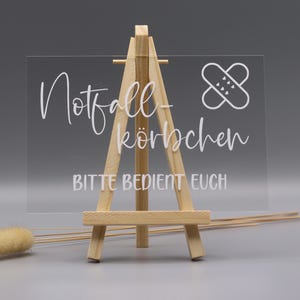 May include: A clear rectangular acrylic sign displayed on a small wooden easel. The sign features white text that reads Notfall-k&ouml;rbchen and BITTE BEDIENT EUCH, accompanied by a white graphic icon of a bandage with small heart details on the surface.