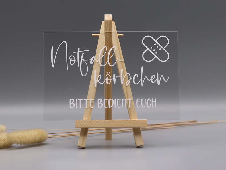 May include: A clear rectangular acrylic sign displayed on a small wooden easel. The sign features white text that reads Notfall-k&ouml;rbchen and BITTE BEDIENT EUCH, accompanied by a white graphic icon of a bandage with small heart details on the surface.