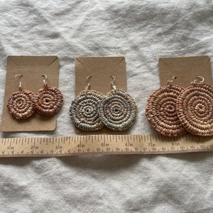 May include: Three pairs of handmade earrings with a circular woven design. The earrings are displayed on brown card stock. The earrings are approximately 5 cm in diameter.