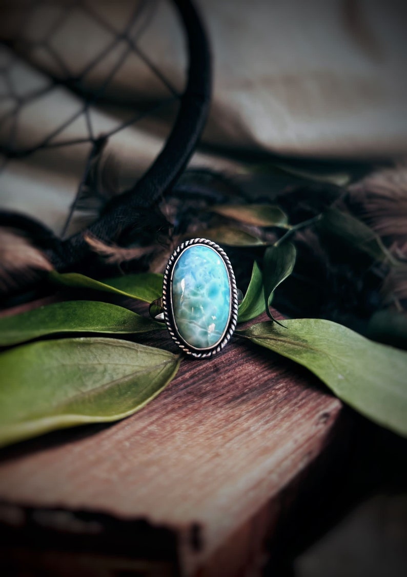 K&ouml;nnte beinhalten: Ovaler Silberring mit einem hellblauen Edelstein, platziert auf einer Holzoberfl&auml;che mit gr&uuml;nen Bl&auml;ttern. Der Stein ist von einem silbernen, seilartigen Rand umgeben. Das Design des Rings strahlt nat&uuml;rliche Sch&ouml;nheit und Eleganz aus.
