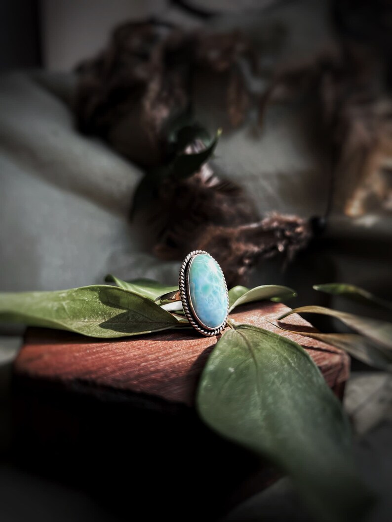 K&ouml;nnte beinhalten: Ein Silberring mit einem ovalen, hellblauen Edelstein. Der Edelstein ist in einer silbernen L&uuml;nette mit einem gedrehten Seildesign gefasst. Der Ring liegt auf einer Holzoberfl&auml;che mit gr&uuml;nen Bl&auml;ttern.