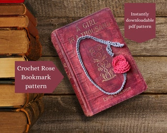 May include: A gray crochet bookmark with a pink crocheted rose, resting on a vintage book titled "A Girl in a Thousand". The book is closed and lying on a wooden surface. The bookmark is a crochet pattern that is instantly downloadable.