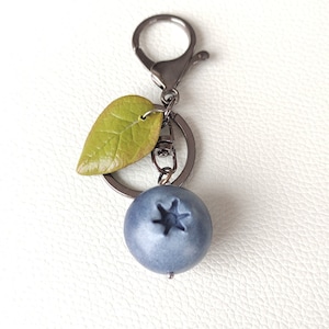 May include: A keychain featuring a round, matte blue blueberry charm with a star-shaped indentation on top, accompanied by a small, textured green leaf charm. The charms are attached to a dark metallic split ring and a lobster claw clasp.