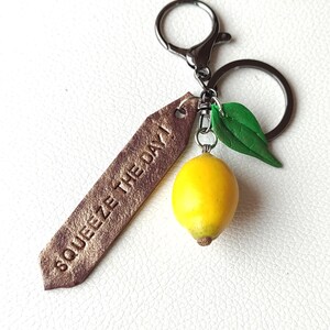 Handmade keychain featuring a realistic lemon charm with a green leaf, attached to a rustic metal ring. A brown, textured tag with the engraved phrase 'Squeeze the Day' adds an inspirational touch, making it a unique and cheerful accessory.