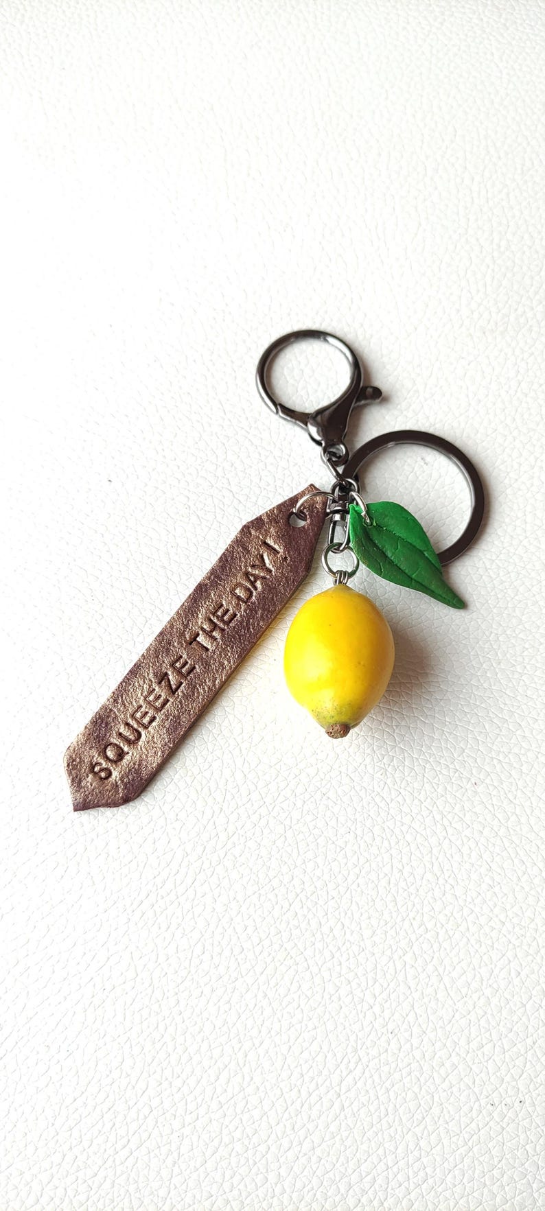 Handmade keychain featuring a realistic lemon charm with a green leaf, attached to a rustic metal ring. A brown, textured tag with the engraved phrase 'Squeeze the Day' adds an inspirational touch, making it a unique and cheerful accessory.