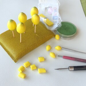 Work-in-progress image of handmade miniature lemons crafted from polymer clay. Small yellow lemons are drying on sticks, surrounded by crafting tools, pigment powders, and tiny lemon slices. A behind-the-scenes look at the handmade creation process.