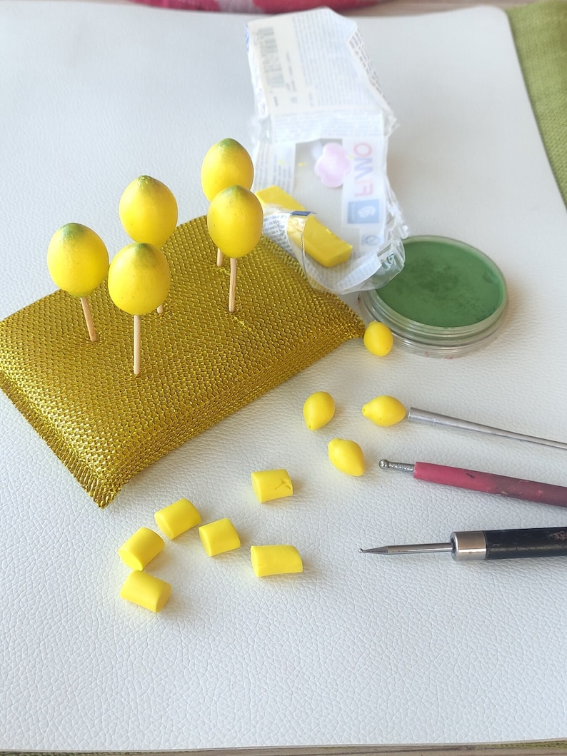Work-in-progress image of handmade miniature lemons crafted from polymer clay. Small yellow lemons are drying on sticks, surrounded by crafting tools, pigment powders, and tiny lemon slices. A behind-the-scenes look at the handmade creation process.