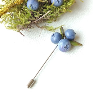 Op de afbeelding: Een zilveren broche met een tros blauwe bosbessen, een populair fruit, op een witte achtergrond.