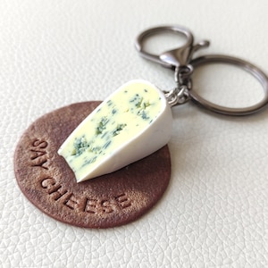 Pode incluir: Um chaveiro de queijo azul com uma casca branca, preso a um chaveiro prateado. O queijo está sobre um círculo de couro marrom com o texto "Say Cheese" em relevo.