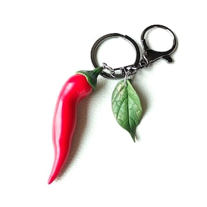 May include: A red chilli pepper keyring with a green leaf charm. The chilli pepper is glossy and detailed, with a green stem. The leaf charm has textured veins. The keyring has a silver-tone clasp and ring.