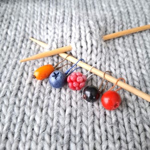 Op de afbeelding: Een set van vijf besvormige steekmarkeerders bevestigd aan een lichtbruine bamboe breinaald die op een grijze gebreide stof rust. De markeerders bevatten een oranje ovaal, een blauwe bosbes, een rode framboos, een zwarte bes en een rode bol.