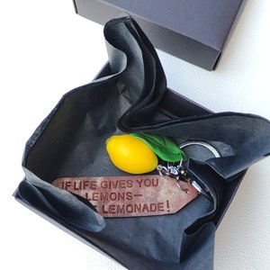 Beautifully packaged handmade keychain with a small yellow lemon charm. Wrapped in black tishue paper and placed inside a gift box. The engraved leatjereffect tag reads 'If life gives you lemons – make lemonade!' A perfect gift for citrus lovers."