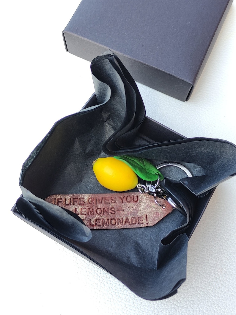 Beautifully packaged handmade keychain with a small yellow lemon charm. Wrapped in black tishue paper and placed inside a gift box. The engraved leatjereffect tag reads 'If life gives you lemons – make lemonade!' A perfect gift for citrus lovers."
