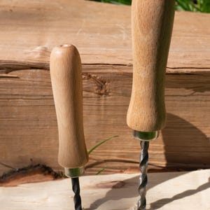 May include: Two wooden handled auger drills, one labeled "Children's" and the other "Adult's". The drills are inserted into a piece of wood. The handles are light brown and the drill bits are black.