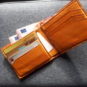 Op de afbeelding: Een bruine leren portemonnee met meerdere kaartsleuven en een geldclip. De portemonnee is open en bevat verschillende creditcards en contant geld. De portemonnee ligt op een grijs stoffen oppervlak.