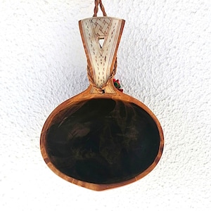 Può includere: Un mestolo di legno fatto a mano con interno scuro e manico decorativo. Il manico presenta un design geometrico di colore chiaro ed è fissato con un cordino intrecciato. Il mestolo è sospeso da un cordino di cuoio marrone.