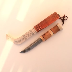 Puede incluir: Dos cuchillos con mangos de madera. El cuchillo de la izquierda tiene un mango blanco con intrincados tallados y una hoja curva. El cuchillo de la derecha tiene un mango marrón con una hoja recta.
