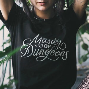 Puede incluir: Camiseta negra con texto blanco en letra cursiva que dice "Master of Dungeons".