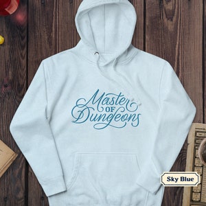 Peut inclure: Un sweat à capuche bleu ciel avec le texte "Master of Dungeons" en écriture stylisée. Le sweat à capuche a une capuche à cordon et une poche avant. L'image est posée sur un fond en bois.
