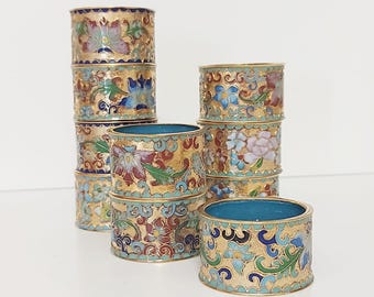 May include: A collection of ornate, gold-toned napkin rings. Each ring features a detailed floral design in shades of blue, red, green, and white, set against a gold background. The interiors are a vibrant turquoise. The rings are stacked and arranged on a white surface.
