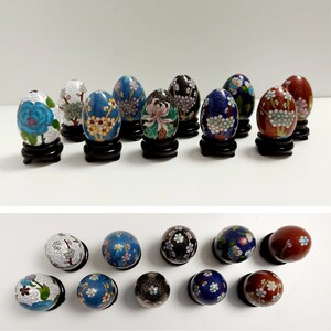 Può includere: Una collezione di 15 piccoli ornamenti a forma di uovo dipinti in modo intricato. Gli ornamenti sono decorati con motivi floreali in tonalità di blu, bianco e rosso. Ogni ornamento è esposto su un piccolo piedistallo in legno nero.