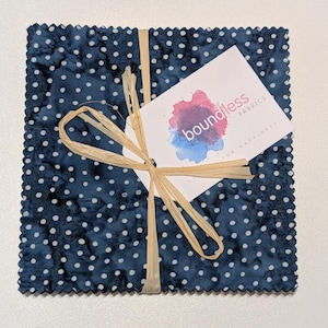 Puede incluir: Una pila de cuadrados de tela azul y blanca con lunares blancos. La tela está atada con un lazo de cuerda natural y una etiqueta blanca con el texto "boundless fabrics" en letras azules.