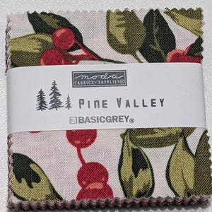 Pode incluir: Um conjunto de quadrados de tecido com um padrão floral de bagas vermelhas e folhas verdes sobre um fundo branco. O tecido é embrulhado com uma etiqueta branca que diz "Pine Valley" e "BasicGrey®". O tecido é da Moda Fabrics Supplies.